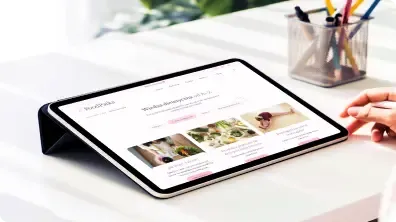 iPad z makietą strony leży na białym biurku, obok kubek z przyborami.