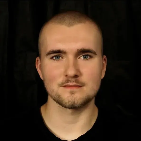 Mężczyzna z zarostem i niebieskimi oczami w czarnej koszulce na czarnym tle.