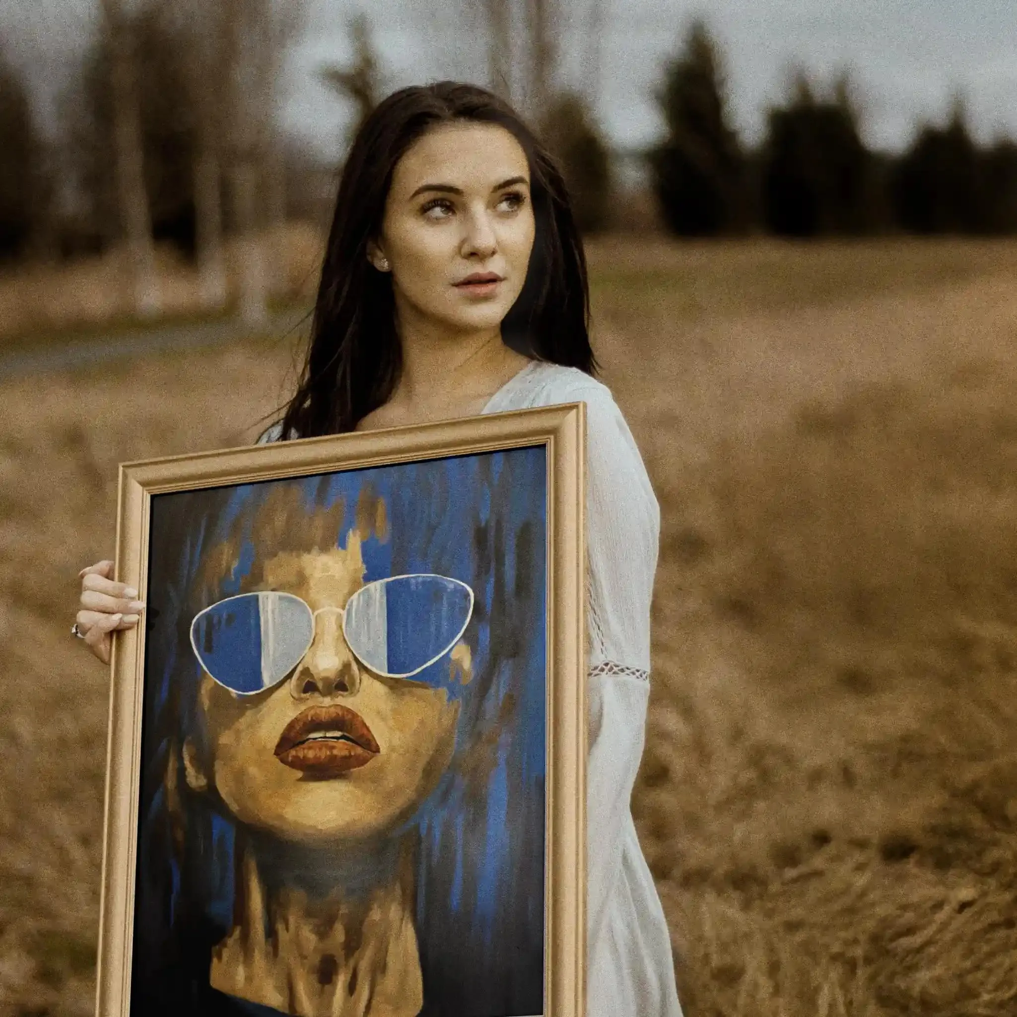 Young woman in a field holds a large portrait of a woman with golden glasses; autumn landscape behind her.