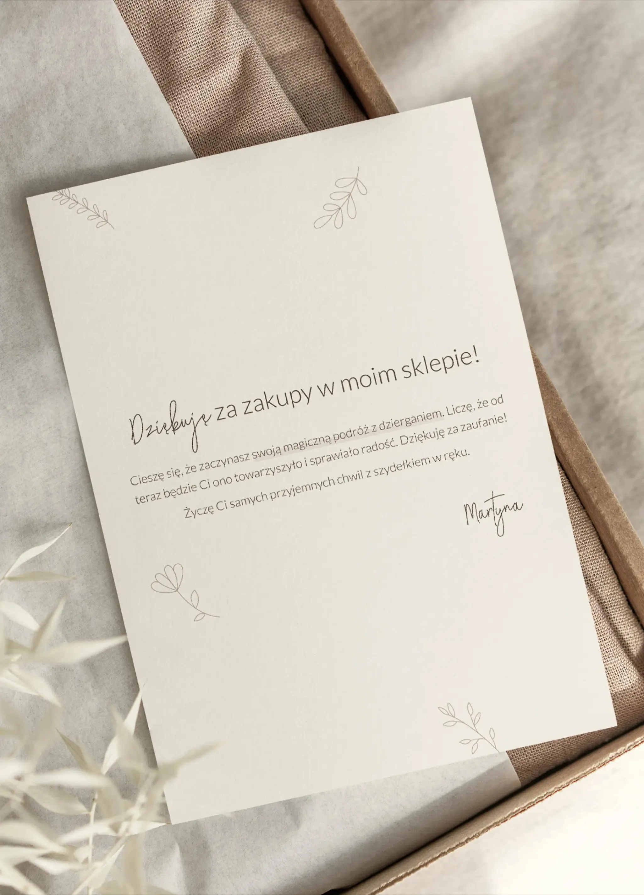 Drukowana kartka z podziękowaniem za zakupy w sklepie, podpisana imieniem właścicielki, Martyna. Papier jest beżowy, leży na tkaninach o delikatnych brązowych i beżowych odcieniach.