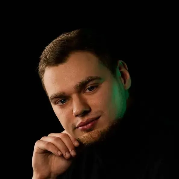 Mężczyzna z brodą opierający się na dłoni, ma czarną bluzkę i stoi na czarnym tle, jego twarz z jednej strony oświetlona jest zielonym światłem.