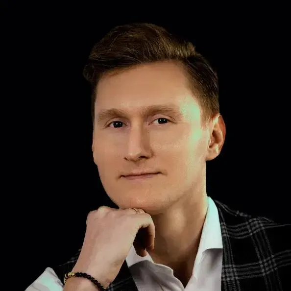 Mężczyzna na czarnym tle, ma na sobie białą koszulę i ciemną kamizelkę w kratę, podpiera się na dłoni, na której znajduje się czarno-złota bransoletka.