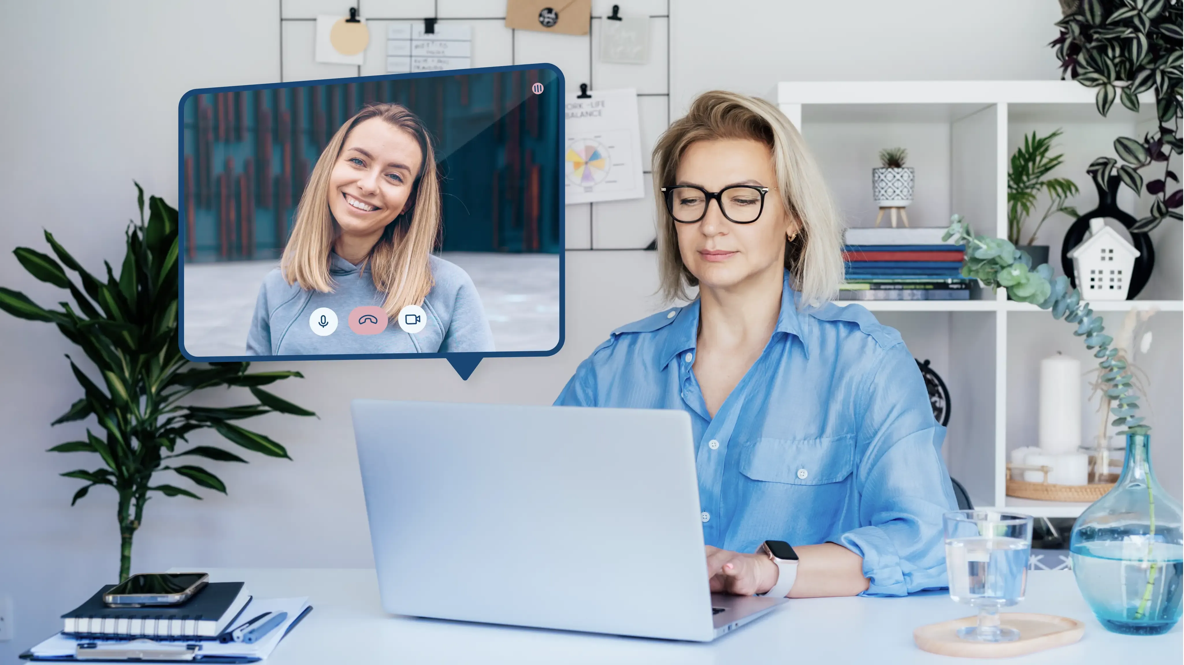 Kobieta w okularach, pracująca na laptopie w domowym biurze, uczestnicząca w wideokonferencji z uśmiechniętą rozmówczynią, której obraz jest wyświetlany na ekranie wirtualnym, otoczona elementami dekoracyjnymi i biurowymi.