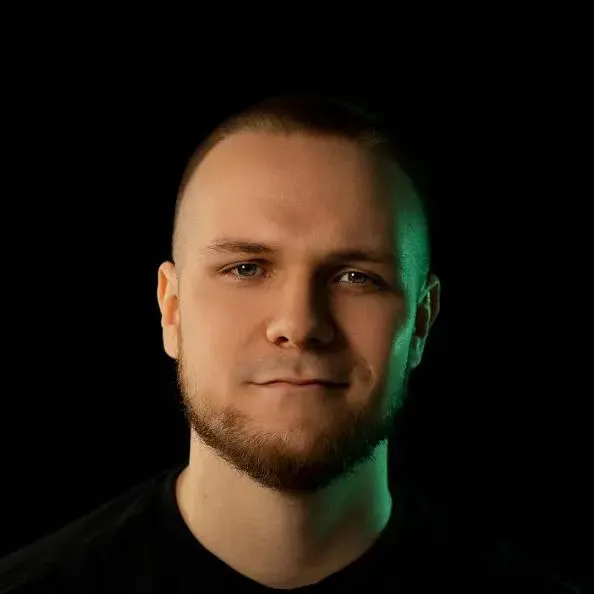 Uśmiechnięty mężczyzna z brodą na czarnym tle i w czarnej koszulce, jego twarz z jednej strony oświetlona jest zielonym światłem.