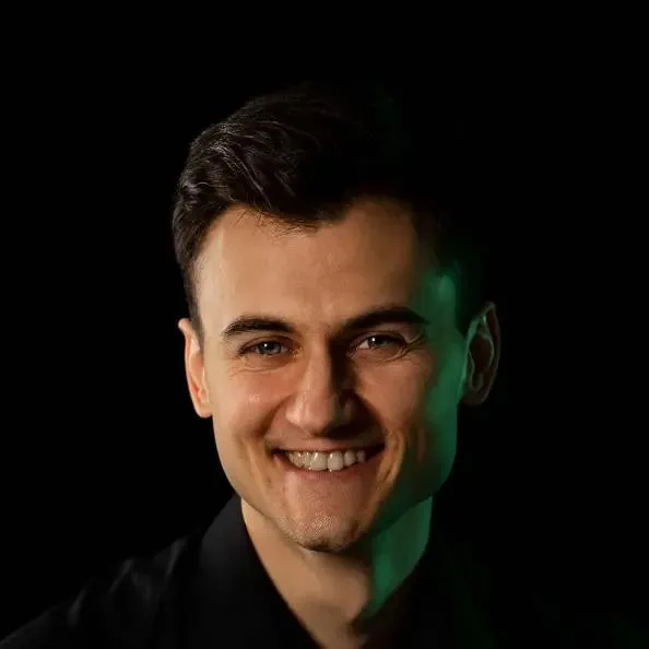 Szeroko uśmiechnięty mężczyzna z ciemnymi włosami, ma na sobie czarną koszulę i stoi na czarnym tle, jego twarz z jednej strony oświetla zielone światło.