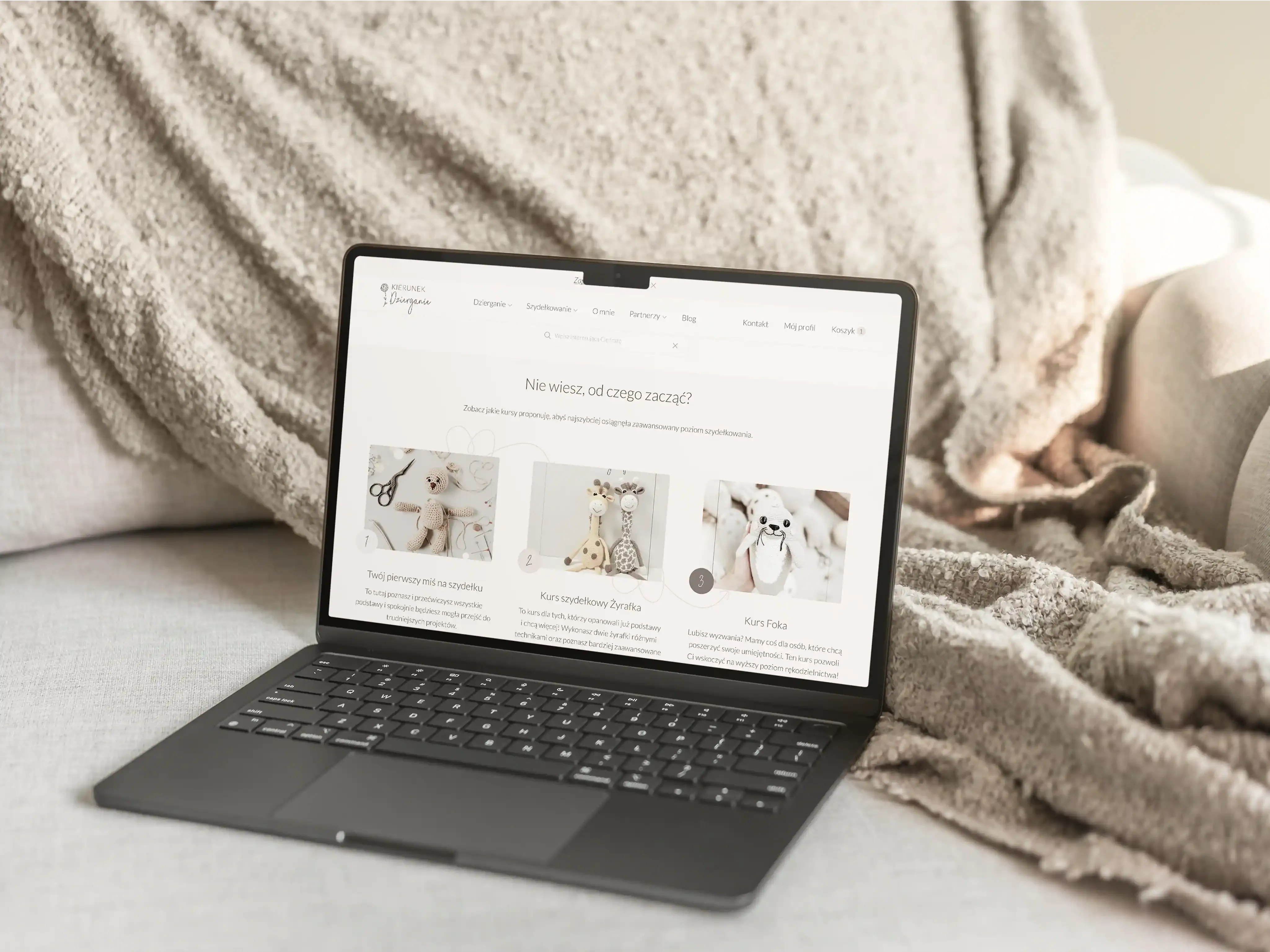 Widok platformy kursowej z kursami z szydełkowania i dziergania na laptopie. Laptop leży na kanapie, w tle beżowy koc o nieregularnej fakturze.